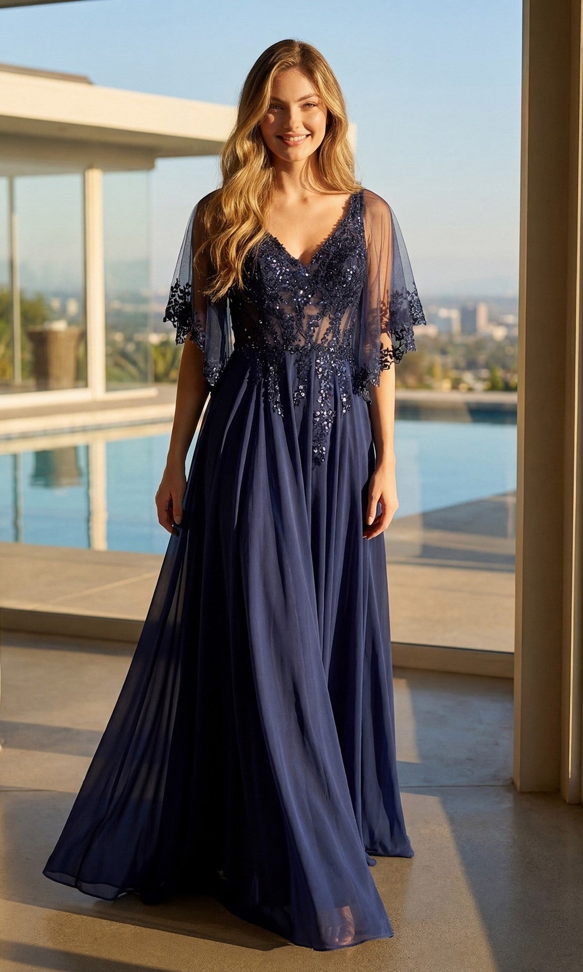 A woman stands indoors by a pool, smiling at the camera, wearing the Juliet JTM22U Long Formal Dress in navy blue, featuring a flowy floor-length a-line skirt and an embroidered sheer bodice.
